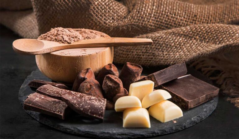 A imagem mostra uma composição elegante de diferentes formas de chocolate sobre uma superfície rústica. Em primeiro plano, há pedaços de chocolate meio amargo, quadradinhos de chocolate branco e blocos de chocolate escuro. Ao lado, vê-se pequenas porções de cacau sólido, em sua forma mais bruta, ressaltando a origem do doce. No centro da cena, uma tigela de madeira contém cacau em pó, com uma colher de madeira apoiada sobre ela. O fundo de tecido de estopa dá um ar artesanal e natural à imagem, remetendo à tradição e ao processo manual da produção do chocolate. A paleta de cores quentes, em tons de marrom e bege, transmite aconchego, sabor e autenticidade. É uma fotografia que convida o leitor a apreciar o chocolate em suas múltiplas versões: do amargo intenso ao branco cremoso, passando pelo cacau em pó, essencial em receitas saudáveis e sofisticadas.