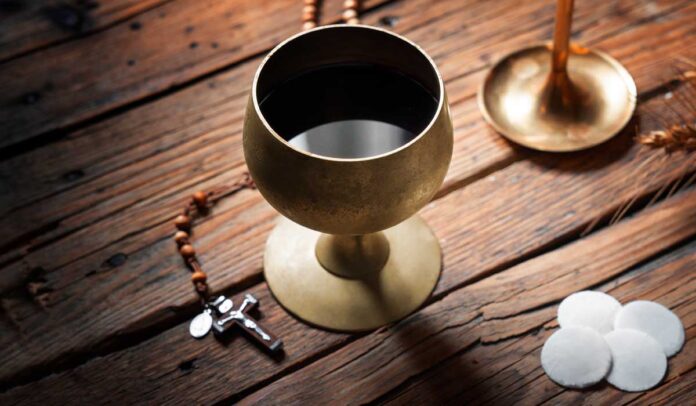 Cálice dourado com vinho tinto ao lado de hóstias brancas tradicionais sobre mesa de madeira rústica, acompanhado de crucifixo, terço e castiçal com vela branca. Imagem simbólica da Eucaristia católica, destacando o vinho como alternativa segura para pessoas com doença celíaca ou restrição ao glúten durante a comunhão.