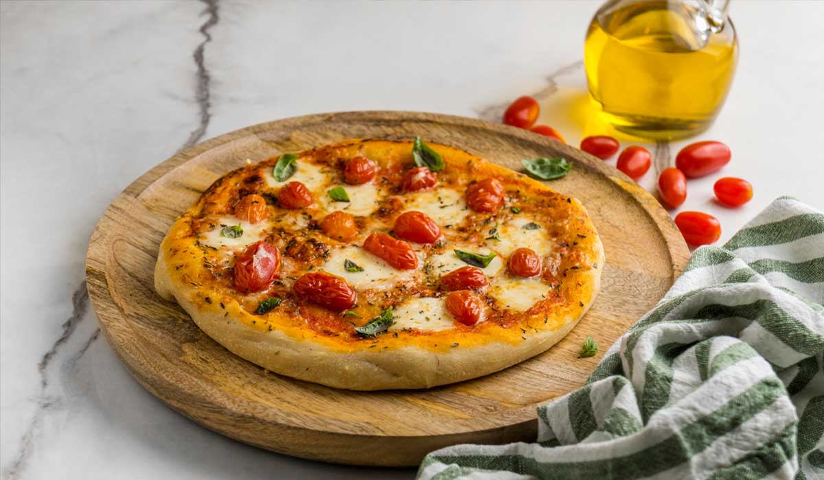Pizza sem glúten estilo margherita, com massa fina e crocante, coberta com molho de tomate, muçarela derretida, tomates-cereja assados e folhas de manjericão fresco. Servida em tábua de madeira rústica, acompanhada de azeite extra virgem e pano de prato listrado verde e branco. Receita 100% sem glúten, perfeita para celíacos que não abrem mão do sabor italiano.
