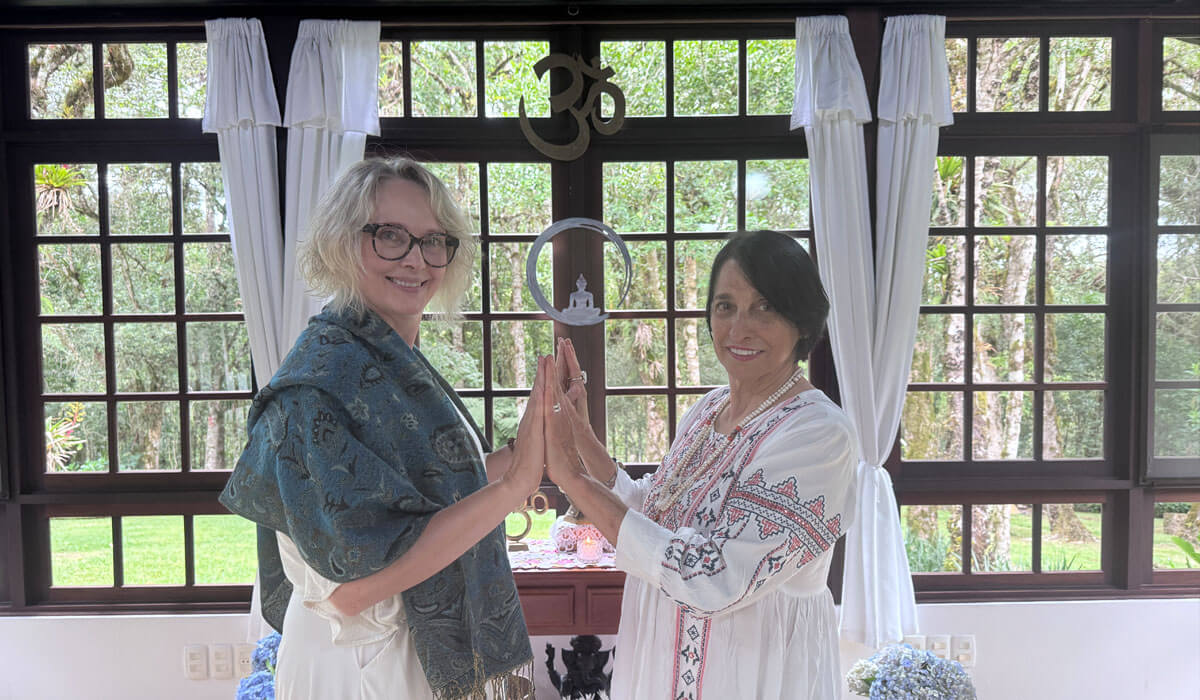 Vanessa Kupczik e Laura Packer em ambiente acolhedor e espiritualizado, com as mãos unidas em gesto de conexão, em frente a grandes janelas com vista para a natureza.