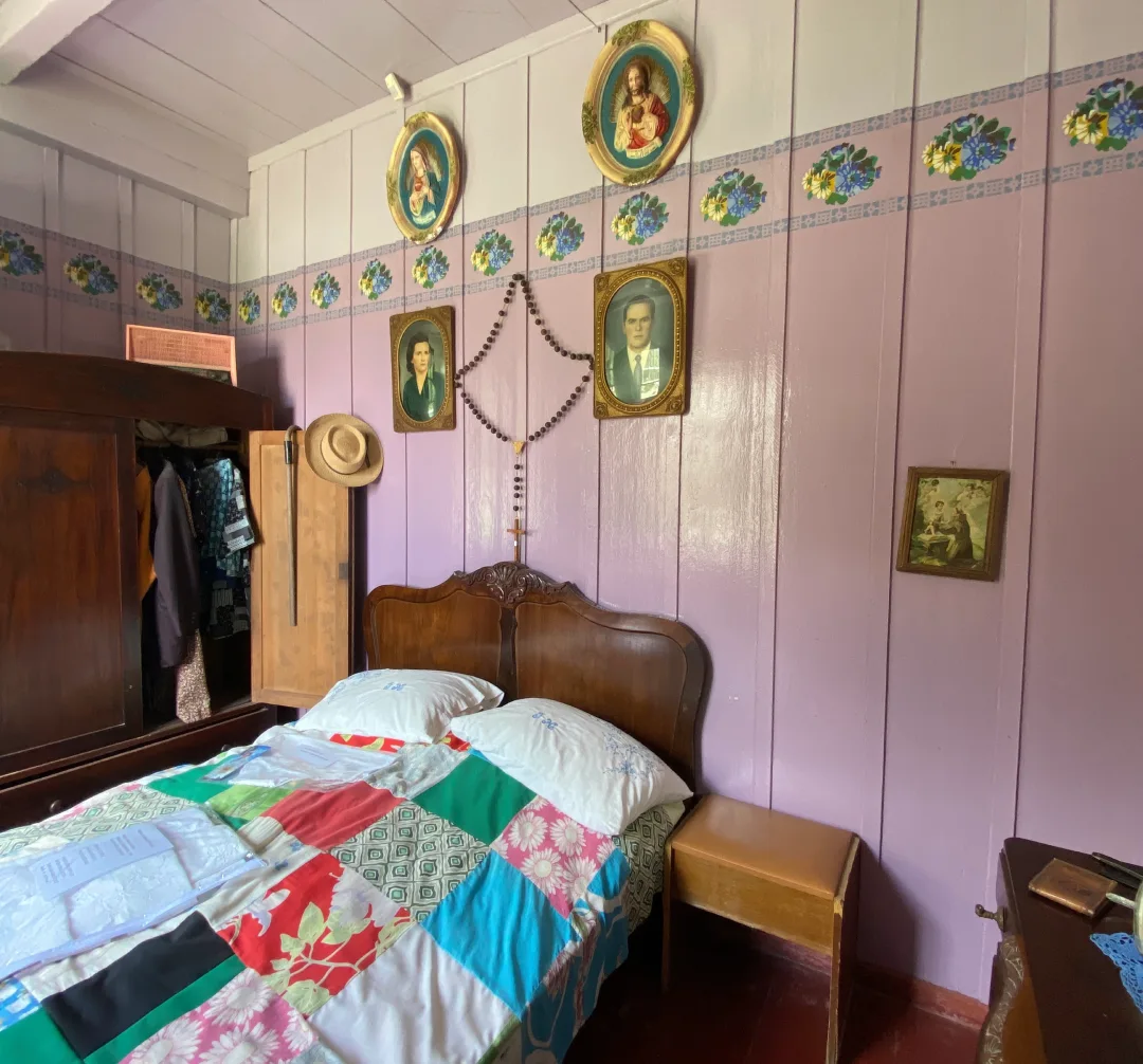 Reprodução de um quarto antigo de uma casa de imigrantes, feito em madeira com paredes em cor de rosa, afrescos e colcha colorida na cama. 